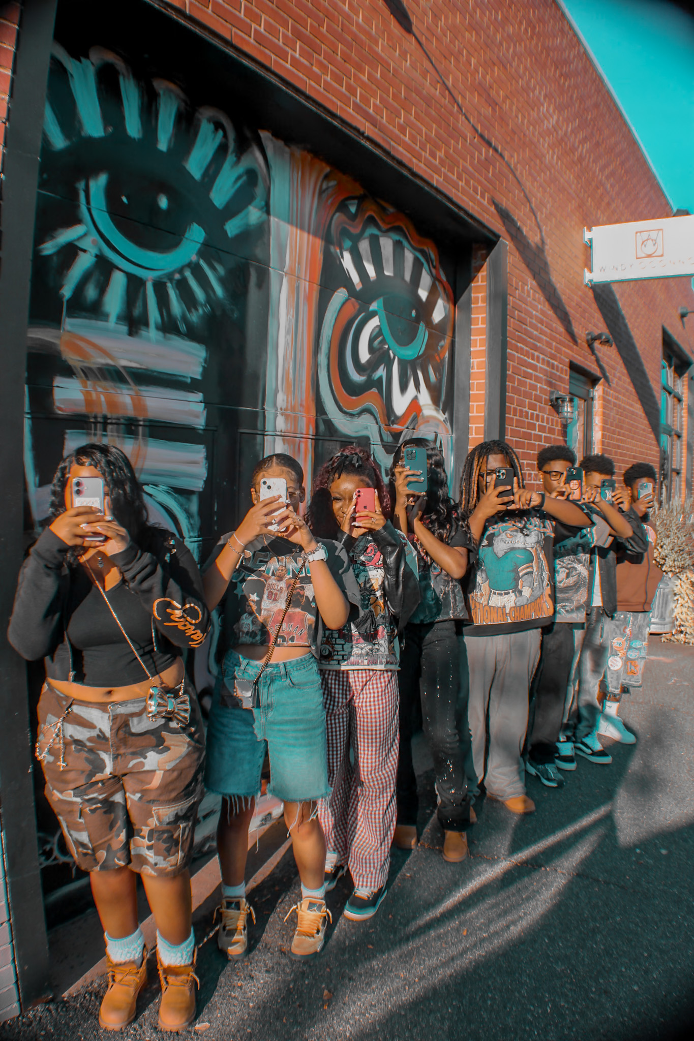 A group of people standing in a line in front of a brick wall mural, each holding a phone in front of their face
